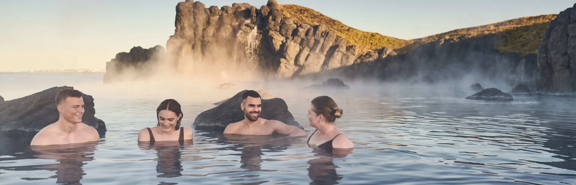 Fyra personer badar i Sky Lagoon bland klippor och vattenånga.
