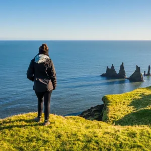 Person på Dyrhólaey med utsikt över Reynisdrangar och Atlanten.